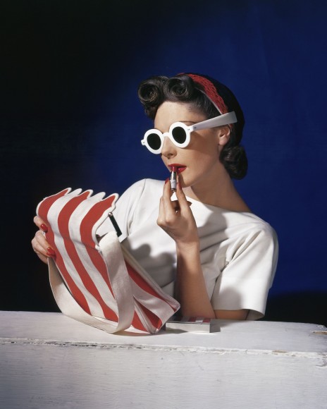Model Muriel Maxwell in white sunglasses putting on lipstick, wearing red-white-and-blue turban, andv holding a red-and-white striped bag